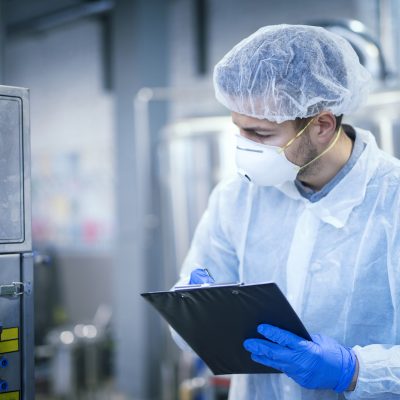 Technologist expert in protective uniform with hairnet and mask taking parameters from industrial machine in food production plant.
